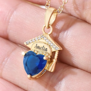 May include: A gold-colored necklace with a pendant shaped like a graduation cap. The cap has the name "Amelia" and is adorned with small, clear crystals. A heart-shaped, deep blue gemstone hangs below the cap.