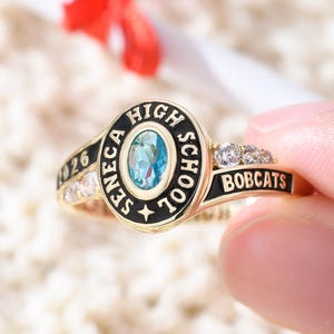 May include: A gold-colored class ring with the words "SENECA HIGH SCHOOL" and "BOBCATS" in black lettering. The ring features a central oval-shaped blue stone and several smaller clear stones. The year "026" is also visible.