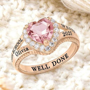 May include: A rose gold ring with a heart-shaped pink gemstone center, surrounded by small clear stones. The ring is engraved with the words "Olivia", "WESTE 2025", and "WELL DONE". The ring is set against a white, textured background.