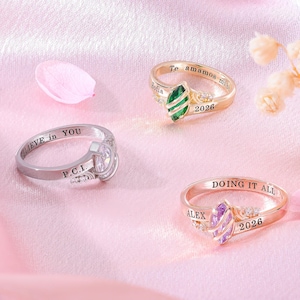 Puede incluir: Tres anillos de metal con piedras de talla marquesa. Un anillo plateado está grabado con "BELIEVE in YOU" y "P.C.I.". Un anillo dorado tiene una piedra verde, "Sofia" y "2026". Un anillo de oro rosa tiene una piedra morada, "ALEX" y "2026".