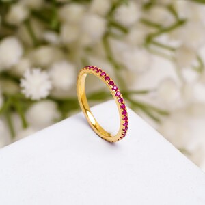 May include: A gold ring featuring a continuous line of small, red gemstones. The ring is set against a white surface with a blurred backdrop of white flowers. The ring's design showcases a band of bright red stones.