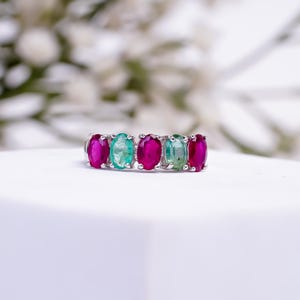 May include: A silver ring featuring five oval-cut gemstones. The gemstones alternate between vibrant fuchsia and light green hues. The ring is set against a soft, blurred background, highlighting the colorful gemstones.