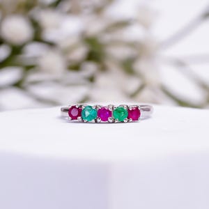 Könnte beinhalten: Ein silberner Ring mit fünf Edelsteinen: zwei rote, zwei grüne und ein magenta. Die Edelsteine sind rund und eng beieinander gesetzt. Der Ring wird auf einer weißen Oberfläche mit verschwommenem floralen Hintergrund gezeigt.