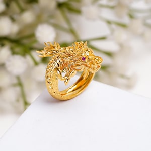 Könnte beinhalten: Ein goldfarbener Ring mit einem detaillierten Drachendesign. Der Drachenkopf ist prominent, mit einem roten Edelsteinauge. Der Ring befindet sich auf einer weißen Oberfläche mit einem verschwommenen floralen Hintergrund, der die komplizierte Handwerkskunst zeigt.