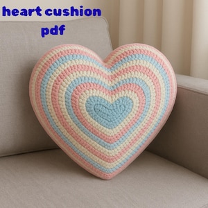 Op de afbeelding: Een hartvormig gehaakt kussen in pastelkleuren roze, blauw en crème. Het kussen heeft concentrische kleurringen, met de tekst "heart cushion pdf" in blauw bovenaan. Het kussen ligt op een grijze bank.
