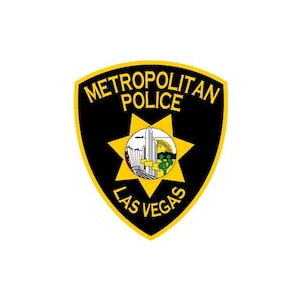 Puede incluir: Un emblema en forma de escudo de la Policía Metropolitana de Las Vegas con fondo negro y borde amarillo. El texto METROPOLITAN POLICE aparece en la parte superior y LAS VEGAS en la inferior. Una estrella amarilla central contiene una escena.
