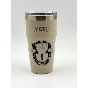 Op de afbeelding: Een beige YETI-beker met een zilveren rand en een doorzichtig deksel. De beker heeft een zwarte afbeelding van een zwaard en pijlen met de woorden "DE OPPRESSO LIBER". Het YETI-logo staat op de voorkant.