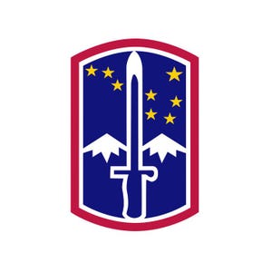 May include: A rectangular patch with a red border, featuring a dark blue field. A white sword stands vertically in the center, overlapping two white mountain peaks. Seven yellow stars are arranged in the upper portion of the blue field.