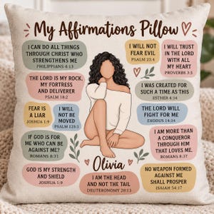 May include: A square throw pillow with a beige fabric cover featuring an illustration of a person with long dark curly hair sitting in the center. The text My Affirmations Pillow is at the top, surrounded by various Bible verses in colored bubbles.