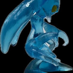 May include: A translucent blue sculpture of a creature with a flowing, amorphous form. The figure has elongated limbs and a head with pointed features. The sculpture has a smooth, glossy texture and is set against a black background.