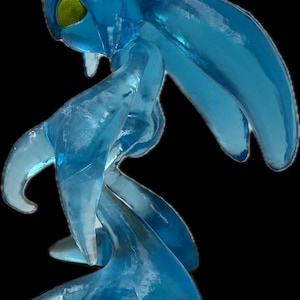 May include: A translucent blue figure with a flowing, amorphous form. The figure has a large head with a yellow eye and a purple marking. Its limbs are elongated and fluid, suggesting movement. The overall design evokes a sense of ethereal beauty.