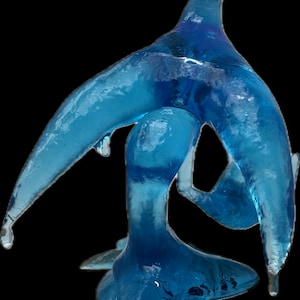 May include: A translucent blue art glass sculpture with an abstract, flowing design. The sculpture has a central, bulbous form with elongated, wing-like extensions and a pointed top. The piece is set against a black background.