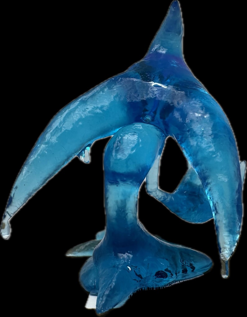 May include: A translucent blue art glass sculpture with an abstract, flowing design. The sculpture has a central, bulbous form with elongated, wing-like extensions and a pointed top. The piece is set against a black background.