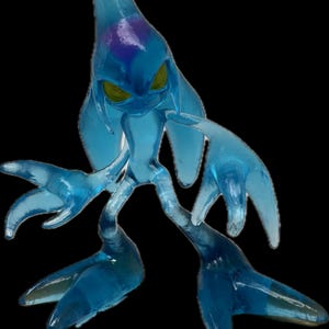 May include: A translucent blue figurine of a creature with a teardrop-shaped head, yellow eyes, and elongated limbs. The figure has a purple area on its head and is posed against a black background. The overall design suggests a fantasy or video game character.
