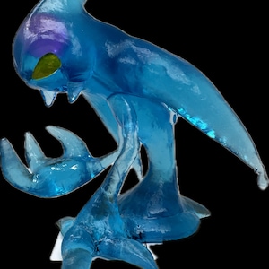 May include: A translucent blue sculpture of a creature with a fin-like head, two arms, and a tail. The sculpture has a smooth, glossy finish with hints of purple and yellow. The creature is posed in a dynamic stance, with its arms outstretched.