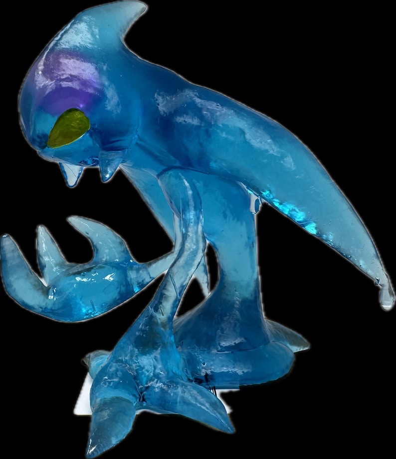 May include: A translucent blue sculpture of a creature with a fin-like head, two arms, and a tail. The sculpture has a smooth, glossy finish with hints of purple and yellow. The creature is posed in a dynamic stance, with its arms outstretched.