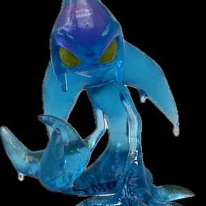 May include: A translucent blue figurine of a creature with a teardrop-shaped head, yellow eyes, and flowing appendages. The figure has a darker blue area on its head and is posed on a black background.