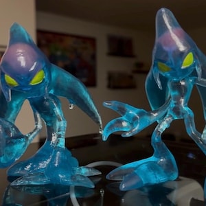 May include: Two translucent blue figures with yellow eyes and a teardrop shape. The figures have elongated limbs and a fluid, watery appearance. The figures are posed on a reflective surface, suggesting a collectible or decorative item.
