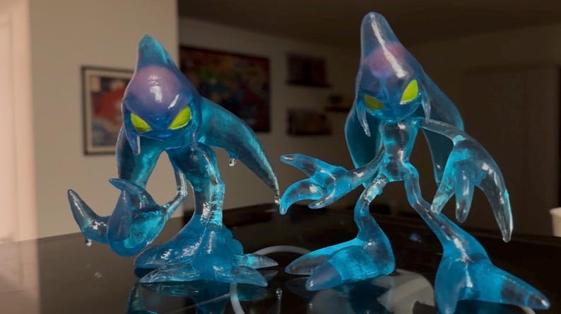May include: Two translucent blue figures with yellow eyes and a teardrop shape. The figures have elongated limbs and a fluid, watery appearance. The figures are posed on a reflective surface, suggesting a collectible or decorative item.