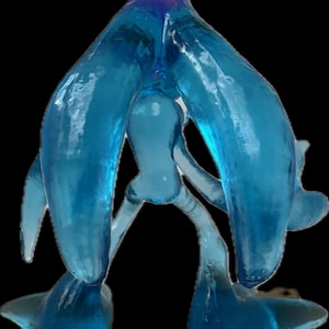 May include: A translucent, light blue art glass sculpture with an abstract design. The sculpture has a central, rounded form with two large, curved arms extending upwards and outwards. The base features two rounded feet, and the overall shape is organic.