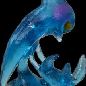 May include: A translucent blue sculpture of a stylized aquatic creature, possibly a dolphin or whale, with a sleek, flowing form. The sculpture has a smooth, glossy finish and is set against a black background.
