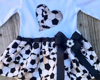 May include: White long-sleeved bodysuit with a heart-shaped soccer ball design. The bodysuit is paired with a black and white soccer ball print skirt with a black bow and a soccer ball button.
