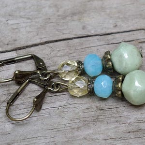 Könnte beinhalten: Ein Paar Ohrhänger mit antiken messingfarbenen Klappbügelverschlüssen. Jeder Ohrring hat facettierte gelbe Perlen, hellblaue Perlen und hellgrüne, abgerundete Perlen. Die Ohrringe werden auf einer verwitterten Holzoberfläche präsentiert.