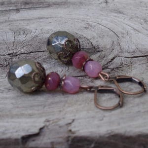 Peut inclure: Une paire de boucles d'oreilles artisanales avec des perles de pyrite facettées, des perles de pierres précieuses roses et violettes, et des crochets d'oreilles cuivrés. Les boucles d'oreilles ont une esthétique vintage.