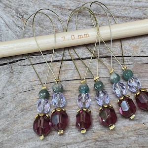 Peut inclure: Ensemble de marqueurs de mailles faits à la main avec des boucles en fil doré. Chaque marqueur présente des perles violettes, vertes et transparentes, avec des accents dorés. Une aiguille à tricoter en bois avec le texte "7.0 mm" est également visible.