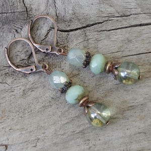 Könnte beinhalten: Ein Paar handgefertigte Ohrringe mit kupferfarbenen Metallhaken und einer Reihe von hellgrünen und klaren Perlen. Die Perlen sind mit kleinen, dunkelbronzenen Metallperlen akzentuiert. Die Ohrringe werden auf einer verwitterten Holzoberfläche präsentiert.
