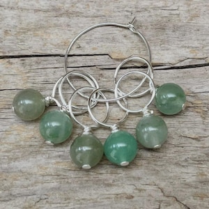 Könnte beinhalten: Silberne Creolen mit sechs grünen Edelsteinperlen. Die Perlen sind rund und haben eine polierte Oberfläche. Die Ohrringe werden auf einer Holzoberfläche präsentiert.