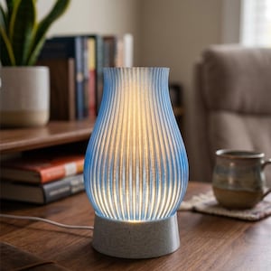 May include: A table lamp with a translucent, ribbed, light blue shade that tapers towards the top. The lamp features a warm internal glow and sits on a speckled, light grey, cylindrical base with a white power cord extending from the side.