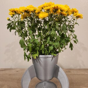 May include: A gray rocket-shaped planter holds a vibrant arrangement of yellow flowers with green foliage. The planter has a cylindrical body, fins, and a circular base. The flowers are in full bloom, creating a striking contrast against the gray planter.