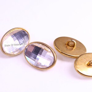 Könnte beinhalten: Vier ovale Knöpfe mit einem facettierten, klaren Kristallmittelteil und einem goldfarbenen Metallrand und -rücken. Die Knöpfe sind auf einer weißen Oberfläche angeordnet. Knöpfe zum Nähen und Basteln.