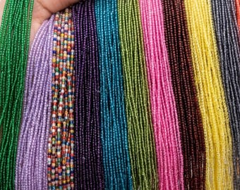 May include: A hand displays strands of colorful seed beads in various colors, including green, purple, blue, pink, yellow, orange, and black. These beads are likely used for jewelry making or other craft projects.