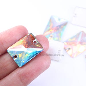 May include: Iridescent rectangular crystal rhinestones with two holes for jewellery making. The crystals reflect a rainbow of colours, including blue, orange, and green. Several crystals are scattered on a white surface.