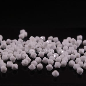 May include: A collection of small, white, faceted beads scattered on a black surface. The beads have a geometric shape with multiple sides and a hole for stringing. The beads are uniform in size and color.