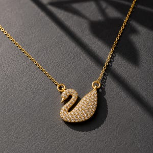 Può includere: Una collana color oro con un ciondolo a forma di cigno. Il ciondolo è incastonato con numerose piccole pietre chiare e scintillanti che ne ricoprono l'intera superficie. Il cigno è fissato a una sottile catenina color oro su entrambi i lati.