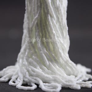 Può includere: Primo piano di un fascio di perline bianche. Le perline sono piccole, rettangolari e infilate insieme, creando un aspetto fluido e strutturato. Il testo "State of Beads Etsy" è visibile. Le perline sono di un bianco brillante.