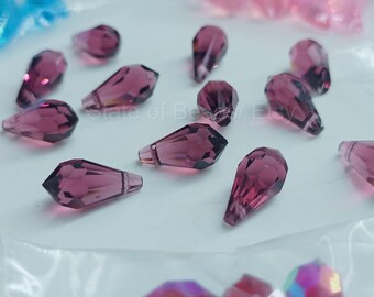 Peut inclure: Gros plan sur plusieurs perles en forme de goutte d'eau, facettées, de couleur magenta foncé. Les perles sont dispersées sur une surface blanche. Ces perles sont probablement destinées à la fabrication de bijoux.