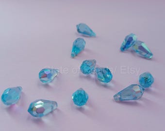 Peut inclure: Plusieurs perles en forme de goutte d'eau bleu clair, facettées, dispersées sur une surface violet pâle. Les perles ont un aspect irisé, reflétant plusieurs couleurs. Idéal pour la création de bijoux.