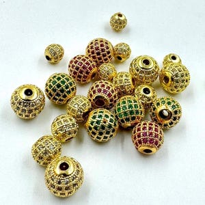 May include: A collection of gold-colored, round beads with intricate detailing. Some beads are adorned with red or green gemstones, while others feature clear, sparkling accents. The beads are scattered on a white surface.