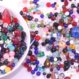 May include: A white bowl filled with a variety of colorful beads, including red, blue, green, and clear. The beads are of various shapes and sizes, including round, faceted, and teardrop. A large red crystal is in the bowl.