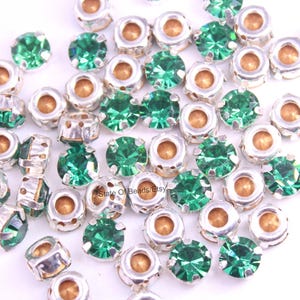 May include: A scattering of small, round, green gemstone beads with silver-coloured metal settings. The beads have a faceted cut, and the settings have a detailed design. The gemstones are scattered across a white surface.