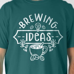 May include: A dark teal short-sleeve t-shirt featuring a white graphic print that reads "BREWING IDEAS" with a lightbulb icon, a coffee cup, coffee beans, and floral accents centered on the chest area of the garment.