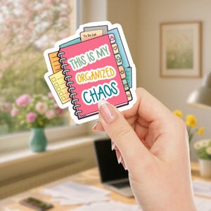 A hand holding a colorful vinyl sticker featuring an illustration of stacked spiral notebooks and planners with tabs. The main text reads 'THIS IS MY ORGANIZED CHAOS' in white, yellow, and teal lettering on a pink notebook cover. The sticker shows various planning elements like to-do lists, checkboxes, and organizational tabs in bright colors including pink, yellow, green, and blue.