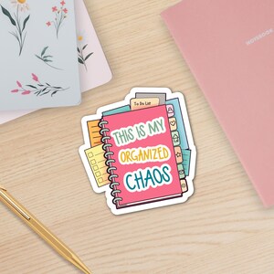 A colorful vinyl sticker featuring an illustration of stacked spiral notebooks and planners with tabs surrounded by Happy Hoos notebooks. The main text reads 'THIS IS MY ORGANIZED CHAOS' in white, yellow, and teal lettering on a pink notebook cover. The sticker shows various planning elements like to-do lists, checkboxes, and organizational tabs in bright colors including pink, yellow, green, and blue.