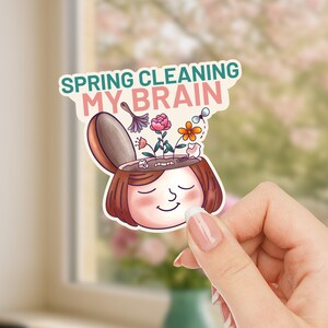 May include: A sticker with the text "SPRING CLEANING MY BRAIN" above a cartoon illustration of a person with an open head, revealing flowers and a cleaning brush. The person has closed eyes and a smiling expression.