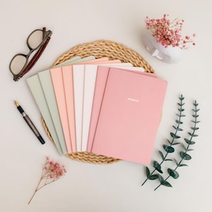 Puede incluir: Una colección de cuadernos en colores pastel, incluyendo rosa, melocotón y verde menta, dispuestos sobre una esterilla tejida. Un par de gafas, un bolígrafo y flores decorativas también están presentes, creando un espacio de trabajo elegante.