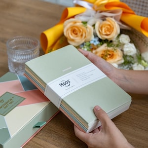 Op de afbeelding: Een cadeau set met een stapel notitieboekjes met een lichtgroene kaft, vastgehouden door handen. De notitieboekjes zijn gebonden met een witte band met de tekst "Hoos". Een geometrisch patroon doos en een boeket oranje rozen zijn ook zichtbaar.